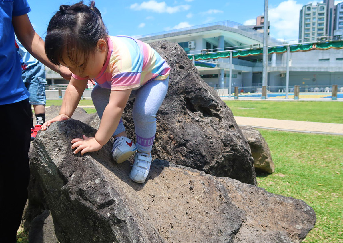 幼兒嘗試攀爬天然岩石，這項非結構化遊戲 (Unstructured Play) 能讓腳底適應不規則表面，刺激前庭覺與本體感覺，訓練真實環境下的平衡適應力。