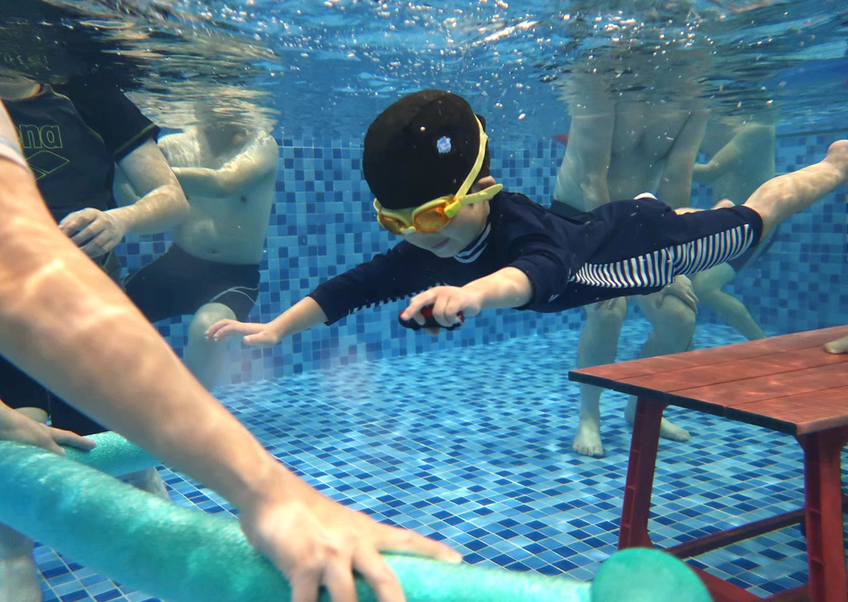 爸爸在水下耐心等待並提供玩具作為視覺目標 (Visual Target)，給予孩子潛水自信。幼兒觀察後主動潛入水中伸展肢體，展現對家長的信任與目標導向的游泳能力。