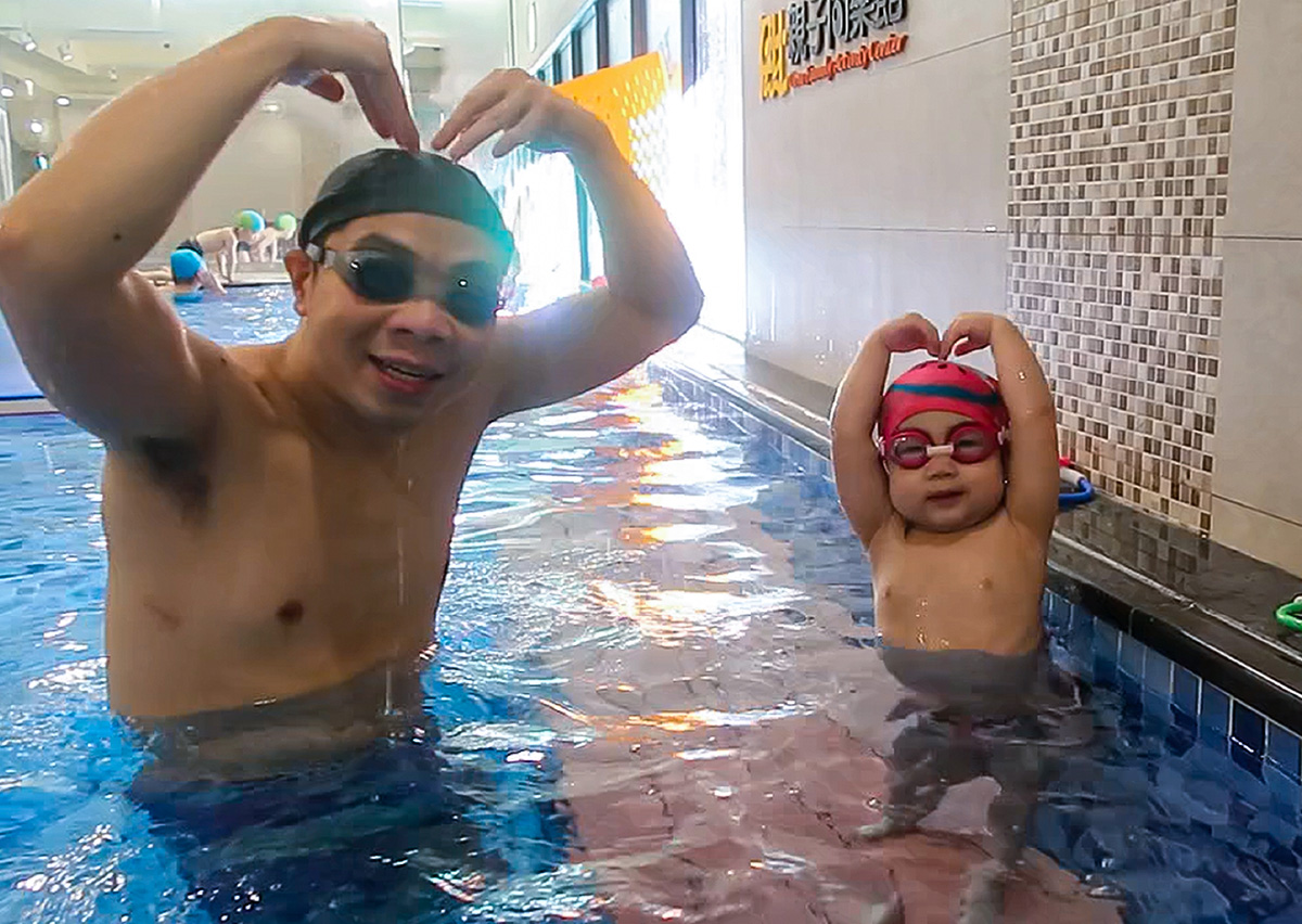 爸爸與孩子在泳池中開心比出愛心手勢 (Heart Gesture)，象徵親子共游帶來的親密情感連結 (Emotional Bonding) 與快樂回憶。透過正向教養與遊戲互動，讓孩子在充滿愛的環境中建立對游泳的熱情與水中自信。