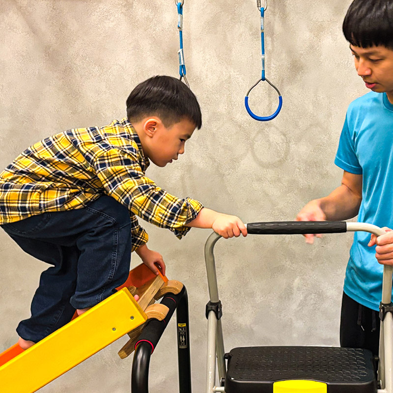 幼兒在教練保護下專注攀爬階梯，這項基本動作技能 (FMS) 訓練能強化動態平衡 (Dynamic Balance) 與下肢肌力，並培養孩子面對高度的專注力。