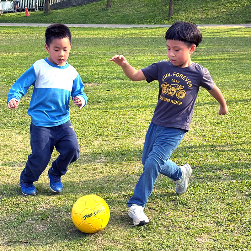 兒童在戶外草地上進行足球奔跑練習，訓練節奏感 (Rhythm) 與物體操控 (Object Control) 能力，這是 FMS 體能課程中關於眼腳協調的重要環節。