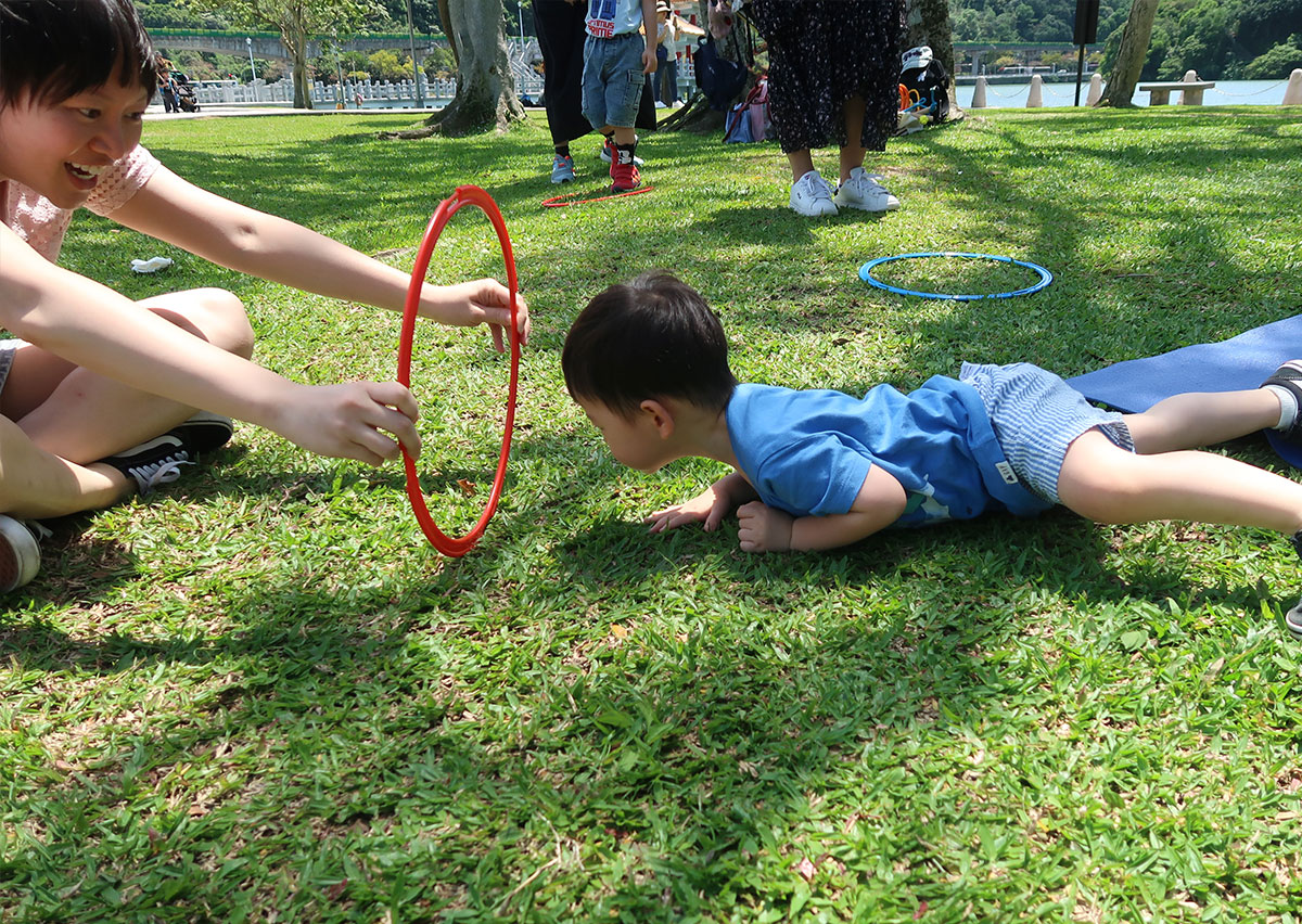 幼兒在戶外草地上進行匍匐爬行穿越呼拉圈，草地的質感能提供豐富的觸覺刺激 (Tactile Stimulation)，爬行動作則能有效強化核心肌群與肩帶穩定度。