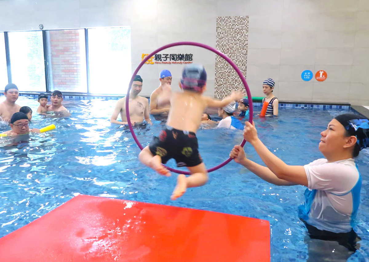 穿著 Oovi 制服的教練手持呼拉圈，引導兒童從浮墊上勇敢跳水 (Jumping Entry) 穿越圓圈，透過遊戲化教學 (Gamification) 訓練入水膽量與空間判斷力。