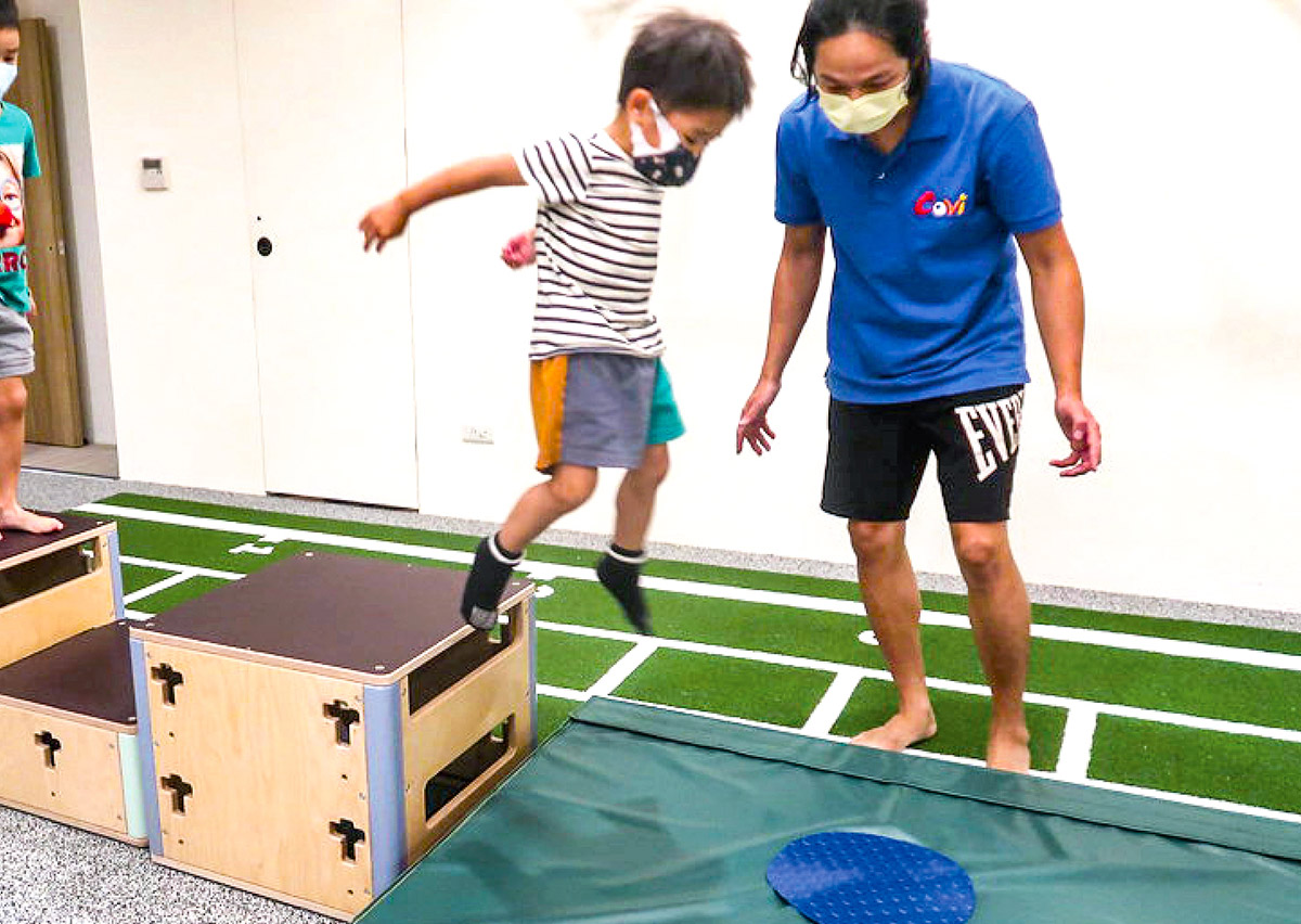 男童從跳箱跳下並練習標準落地緩衝 (Safe Landing) 姿勢，Oovi 教練在一旁隨時保護。這項訓練教導孩子如何利用肌肉吸收衝擊力，保護膝蓋腳踝，是預防運動傷害的關鍵課程。