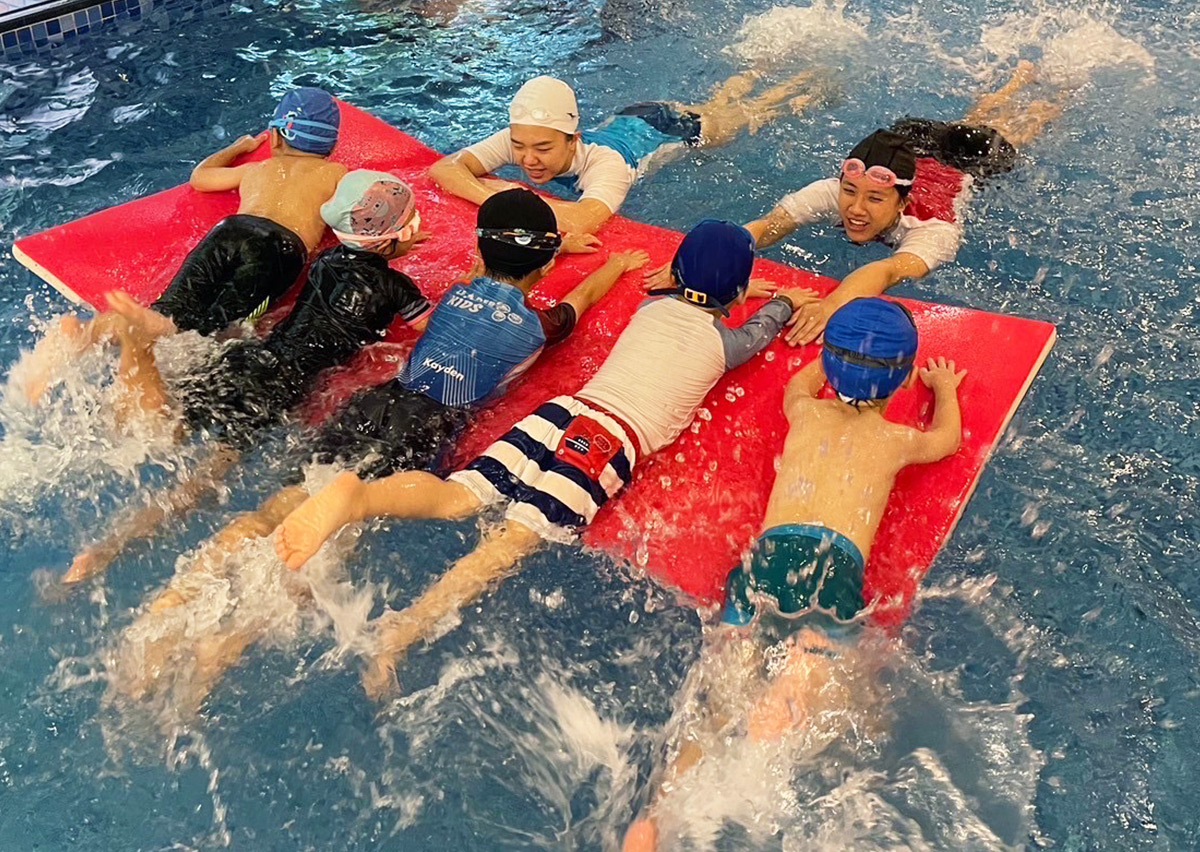 Oovi 專業教練指導兒童趴在浮墊上進行打水練習 (Kicking),透過團體訓練強化腿部肌力與腳踝柔軟度,並幫助孩子記憶游泳時的水平身體流線 (Streamline)。