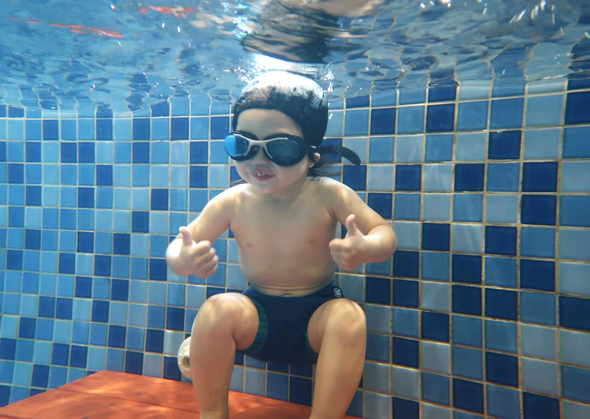 兒童在水中自在坐在長凳上，配戴泳鏡並比出雙讚手勢 (Thumbs Up)，展示極佳的水中自信 (Water Confidence) 與熟練的閉氣控制能力，象徵快樂的學習成果。