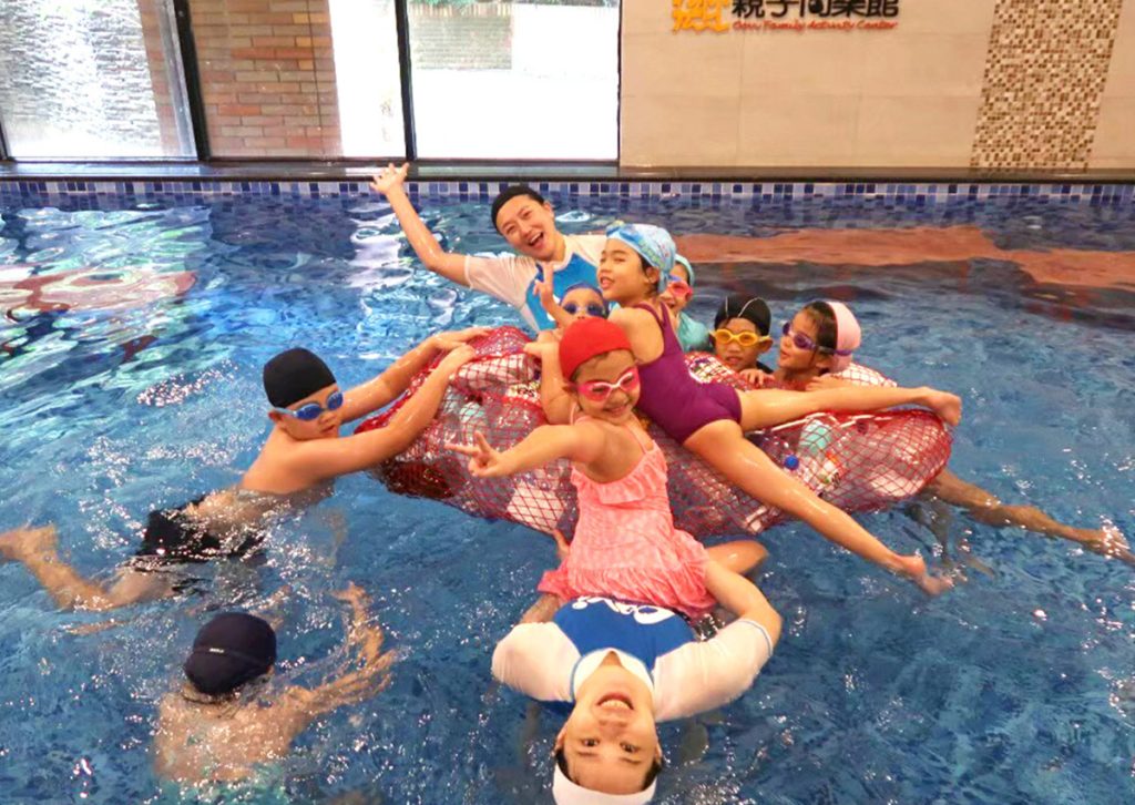 穿著 Oovi 制服的專業教練帶領兒童進行「山大王」水中浮墊挑戰遊戲，透過遊戲化學習 (Gamification) 訓練平衡感，並在快樂氛圍中促進同儕間的社交能力與團隊合作。