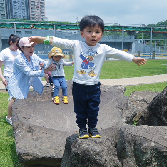 兒童在戶外進行自然探索與體能訓練，雙手張開站在岩石上維持動態平衡 (Dynamic Balance)。這項練習能強化核心肌群與本體感覺，並在克服高度恐懼的過程中建立強大的自信心。