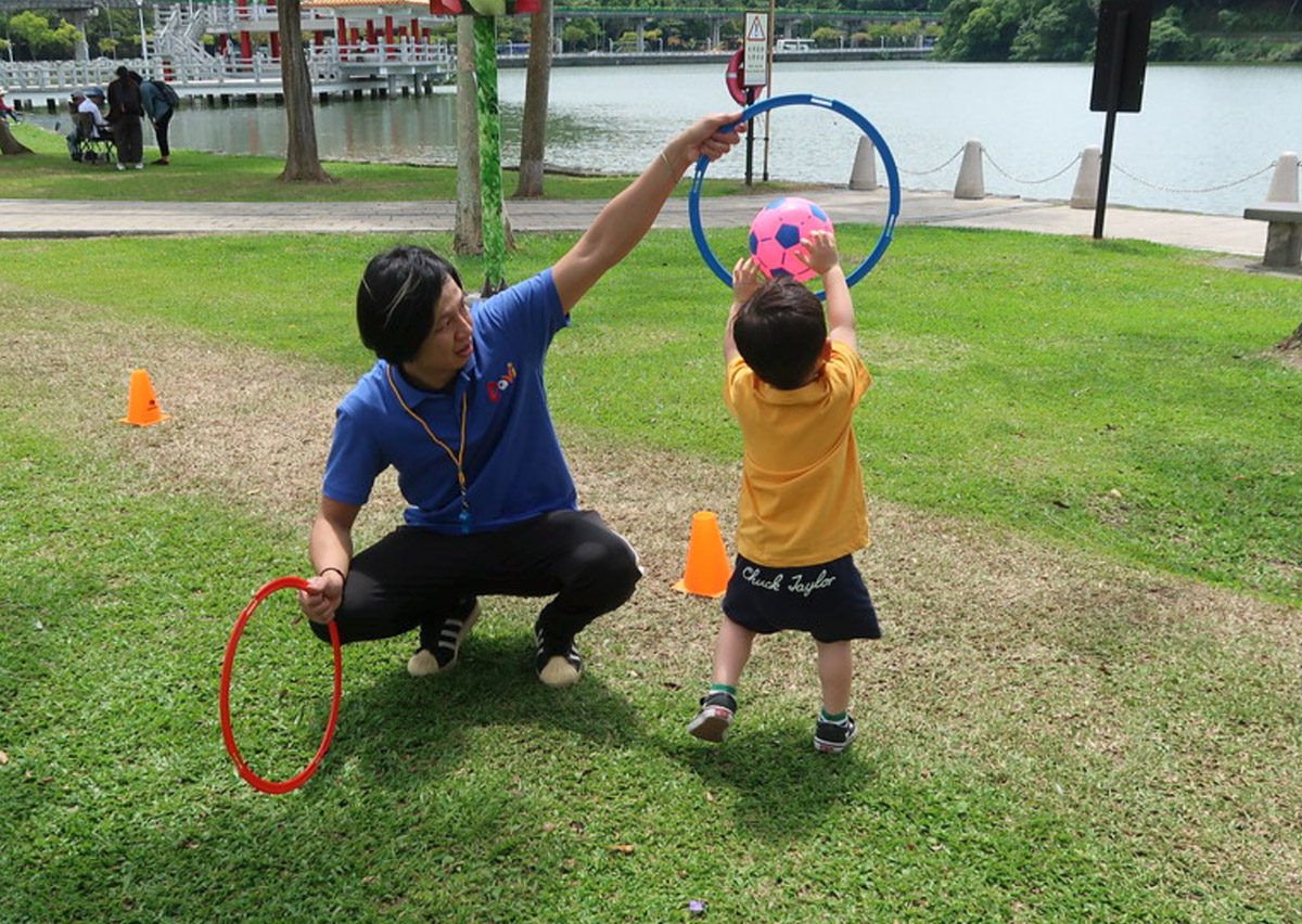 教練在戶外手持圓環作為目標，幼兒練習將球投入圈中，這項活動專注於訓練物體操控 (Object Control) 與空間距離判斷，強化手眼協調能力。