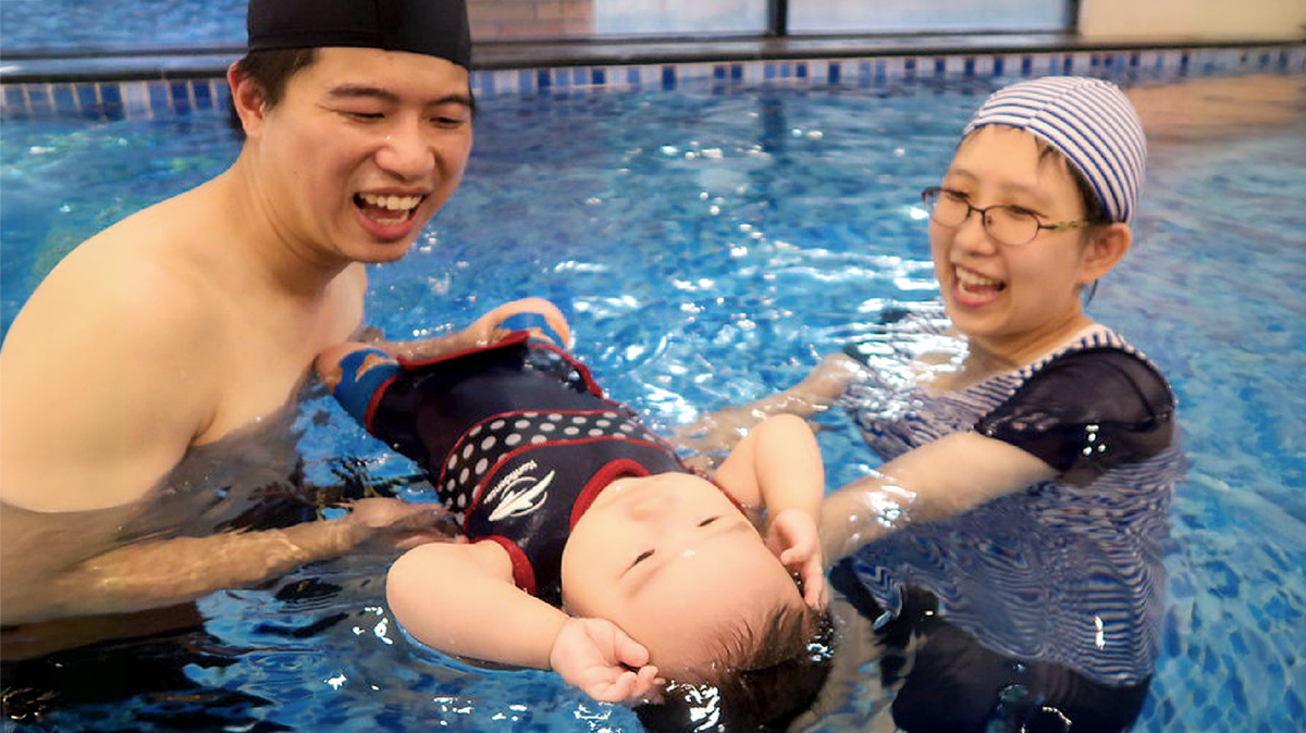 父目前雙手溫柔托住穿著防寒衣的嬰兒進行仰漂 (Back Float) 練習，教練在旁細心指導，建立寶寶對水的信任感並促進親子親密依附關係。