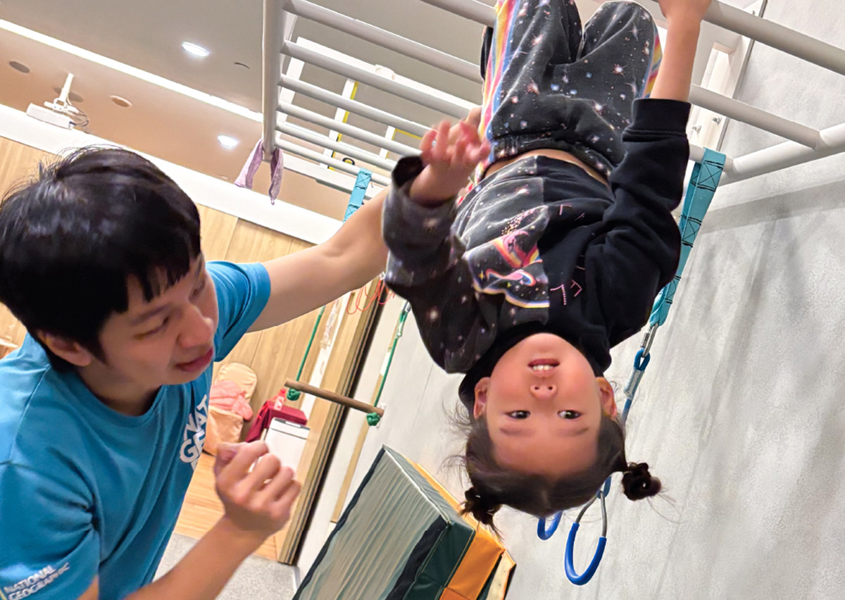 女童在教練保護下進行單槓倒掛練習，透過頭部位置改變強烈刺激前庭覺系統 (Vestibular System)，幫助大腦整合空間方位感，提升孩子的抗暈眩能力與自信。