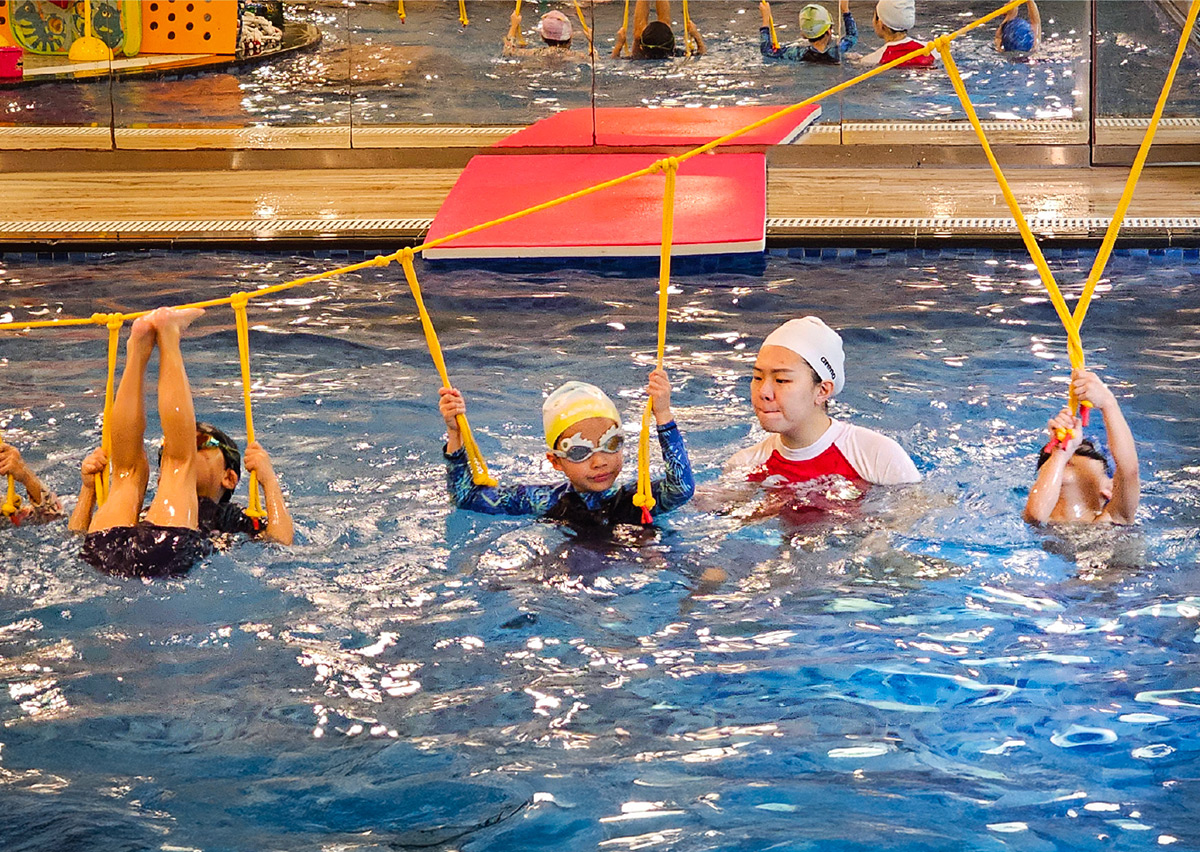 兒童在教練看護下挑戰水上黃色繩索懸吊,進行翻轉與攀爬動作,這項趣味活動能有效訓練上肢肌力 (Upper Body Strength) 與核心協調性,同時培養水中膽量。