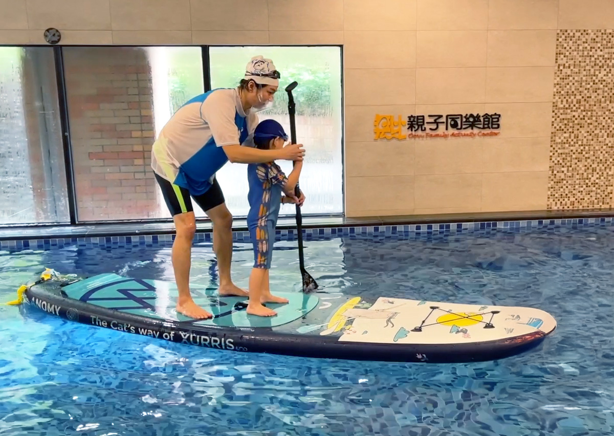 穿著 Oovi 制服的專業教練指導兒童在室內泳池進行水上立槳 (SUP) 訓練,引導孩子在不穩定的板上維持動態平衡 (Dynamic Balance) 並操作划槳。這項特色課程能同時強化核心肌群、專注力與全身協調性。