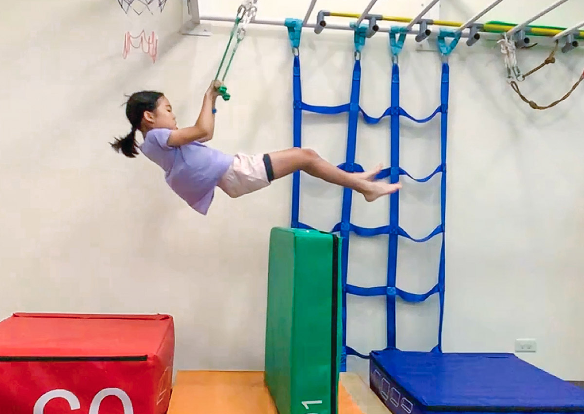 女童利用懸吊握把擺盪穿越障礙物，這項動作需要強大的上肢抓握力與核心收縮能力，訓練身體在空中的動態控制與節奏感。