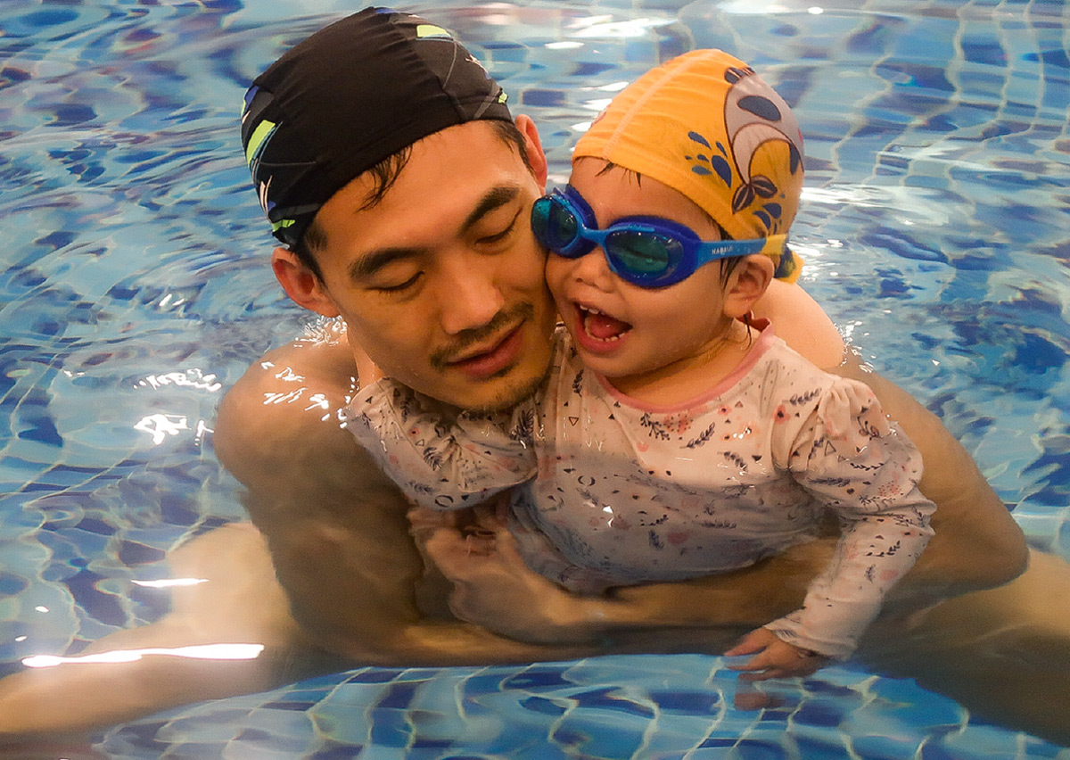 爸爸抱著配戴泳鏡的幼兒在水中親密互動，透過正向鼓勵讓孩子習慣穿戴游泳裝備，並享受親子共游的快樂時光。