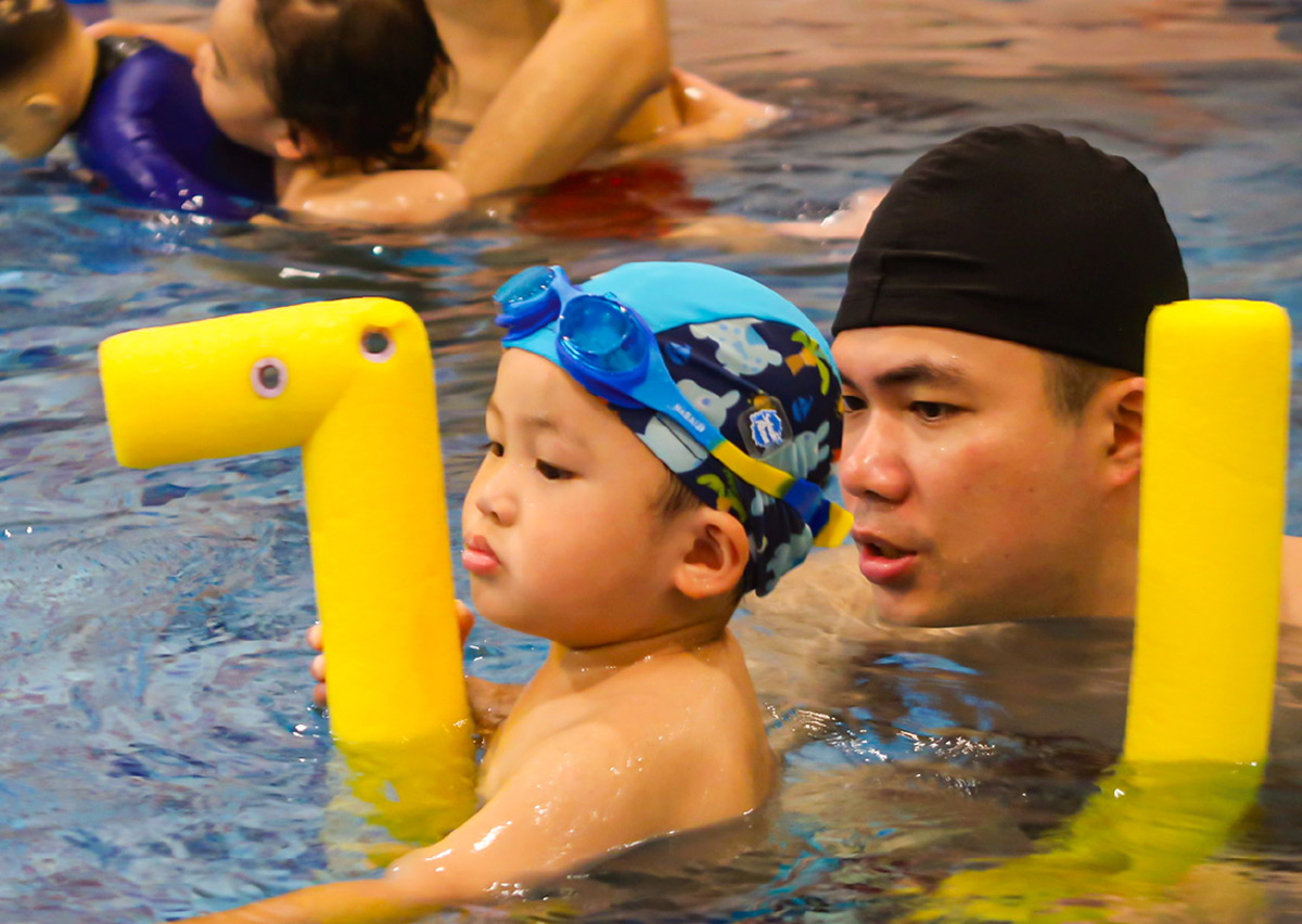 爸爸陪伴幼兒使用黃色浮條進行打水練習，透過教具輔助建立水中浮力認知，並強化親子共游的信任感與安全感。