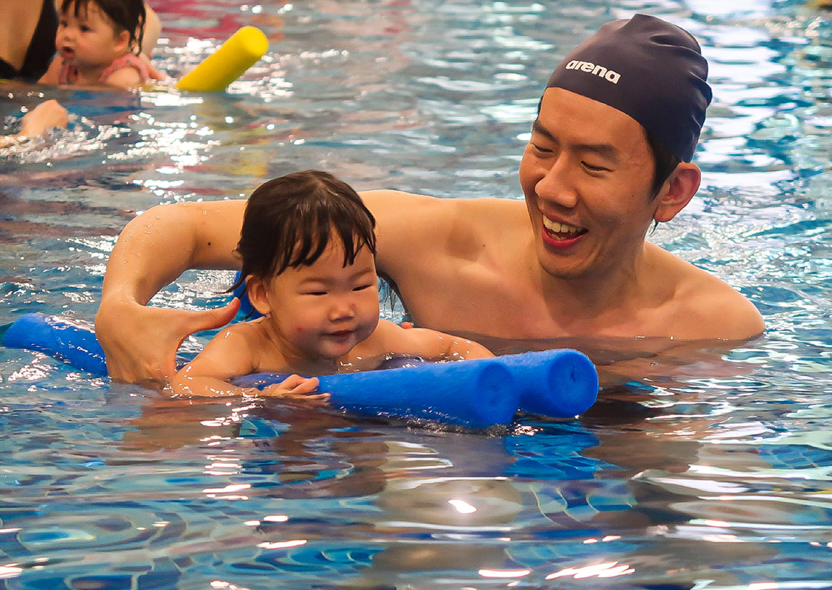 爸爸與孩子在泳池中使用藍色浮條互動大笑，透過遊戲化學習 (Play-based Learning) 讓孩子對游泳產生正向連結，降低學習壓力。