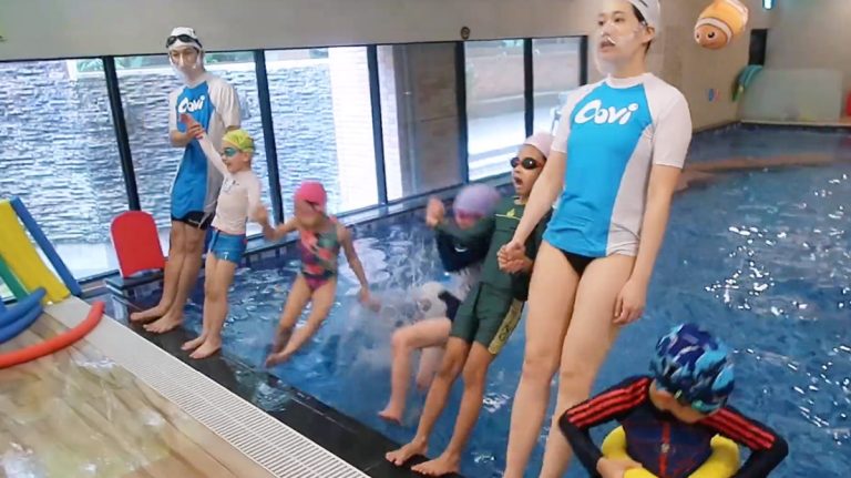 穿著 Oovi 制服的專業教練在岸邊牽著孩子的手,引導學員排隊練習跳水入池。這項勇氣訓練旨在透過安全輔助與團體激勵,幫助兒童克服怕水恐懼 (Overcoming Water Fear) 並建立入水膽量。