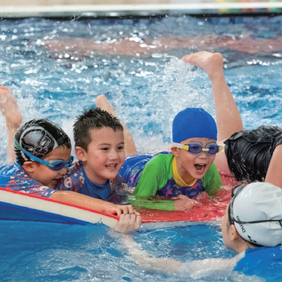 一群兒童趴在大型浮墊上進行團體打水練習 (Kicking)，透過製造水花的趣味競賽訓練腿部肌力，並在歡樂氛圍中促進同儕間的社交互動 (Social Interaction)。