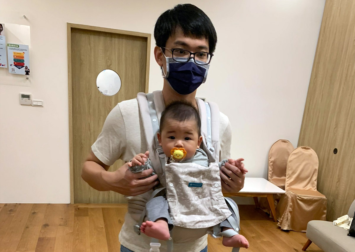 爸爸使用插扣式揹巾 (SSC) 面外揹寶寶，寶寶腿部呈現標準的 M字腿 (M-Shape) 健康坐姿，保護髖關節發育。這張照片鼓勵爸爸參與育兒，面外揹讓寶寶探索。