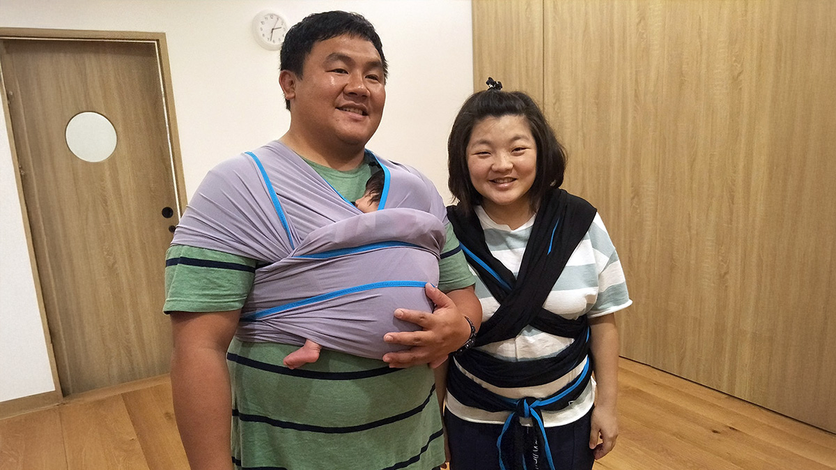 爸爸自信地使用揹巾揹著寶寶，與媽媽開心合影。強調爸爸參與 (Father Involvement) 育兒能有效分擔媽媽辛勞，並透過貼身接觸建立深厚的父嬰連結 (Father-Infant Bonding)。