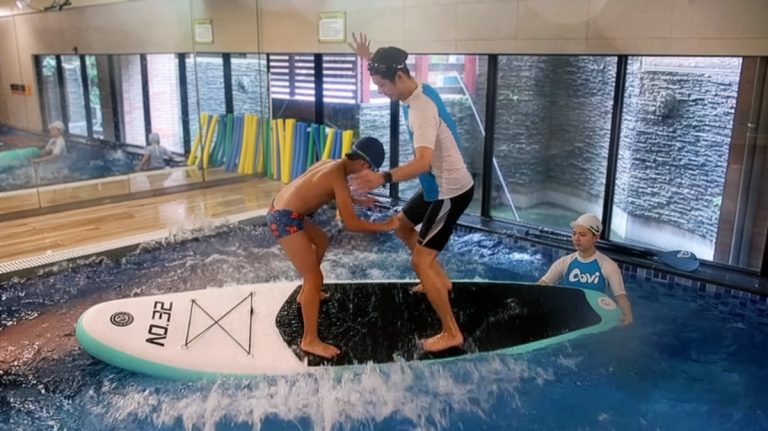 兒童在 Oovi 教練保護下挑戰水上 SUP 立槳板，在不穩定的水面上練習站立，這項進階挑戰能深度刺激前庭覺並強化全身核心肌群 (Core Stability)。