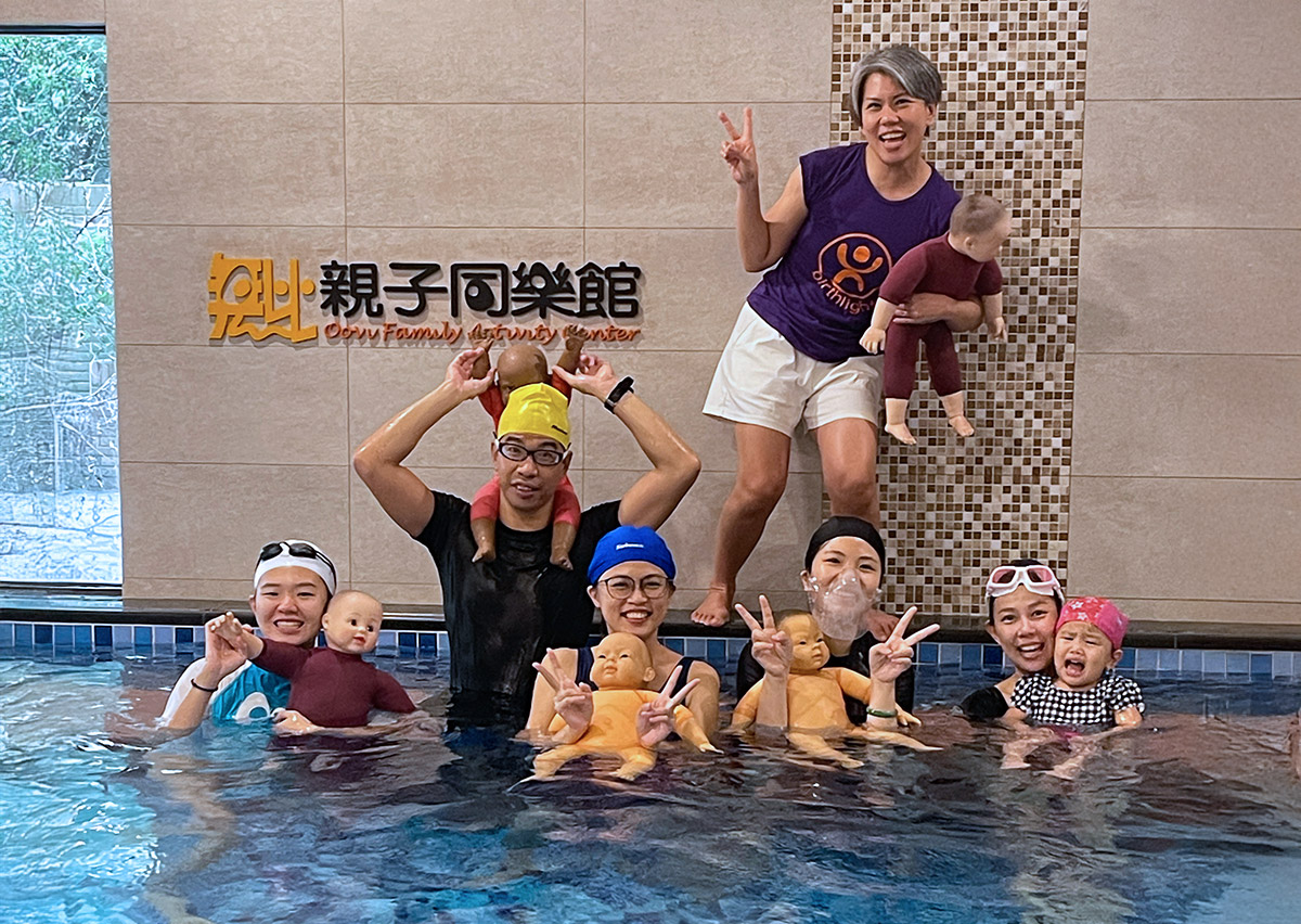Oovi 親子同樂館專業教練團與培訓學員在泳池內開心合影，背景為 Oovi Family Activity Center 標誌。教練們使用專業教具娃娃進行水中模擬練習，展示 Birthlight 英國生之光 國際認證師資的嚴謹培訓流程與團隊熱忱。