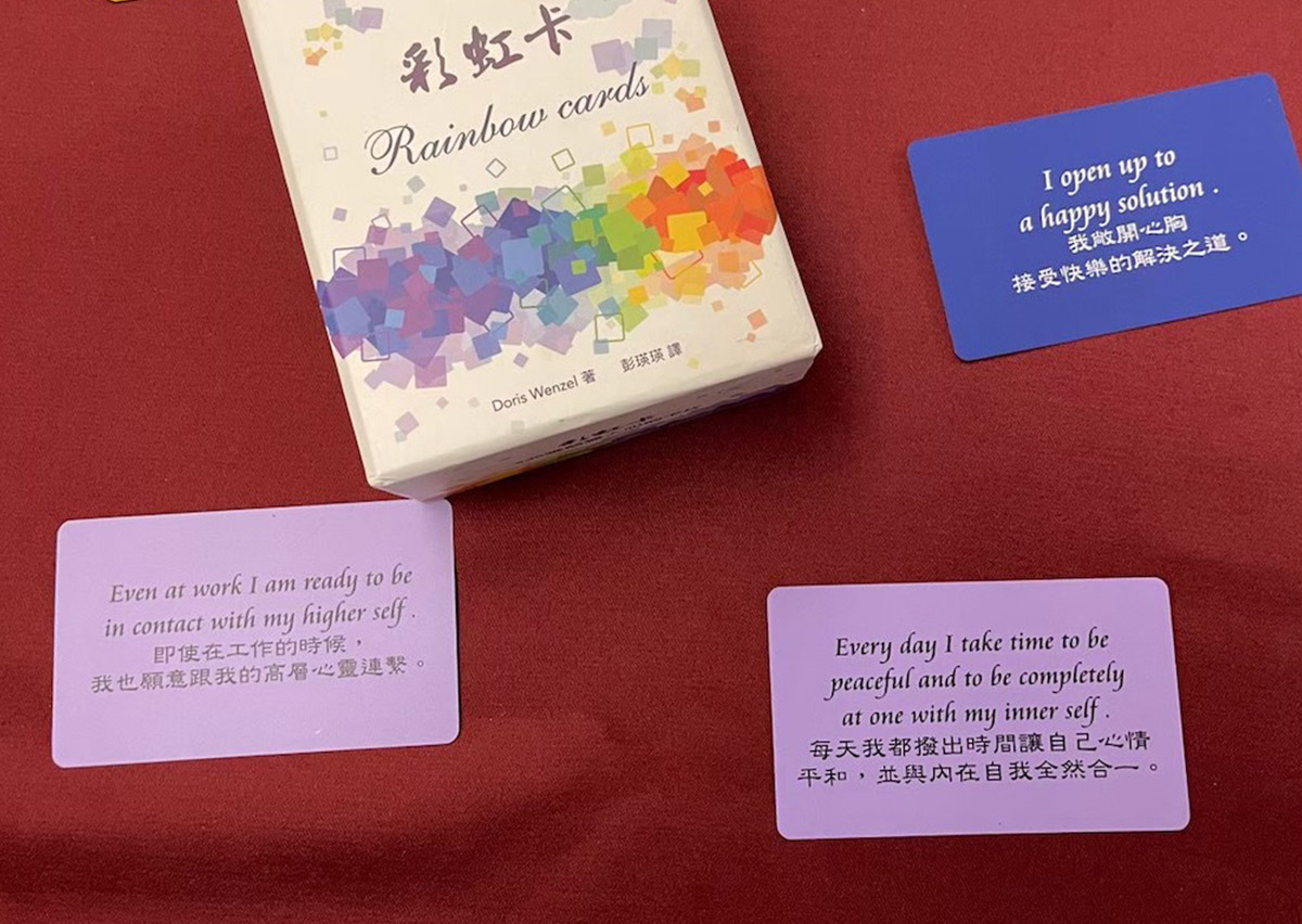 Oovi 揹巾穿戴師資培訓：彩虹卡 (Rainbow Cards) 與內在指引同學分享。「彩虹卡 (Rainbow Cards)」外盒與三張示例卡片陳列於紅色桌面上。卡片上印有中英文對照的心靈指引短句，例如藍色卡片上的「我敞開心胸接受快樂的解決之道」，用於輔助學員進行內在覺察與情緒引導。