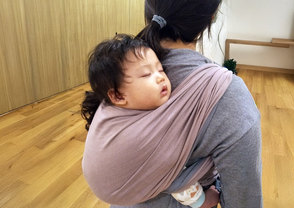 幼兒在媽媽背上使用揹巾進行後揹 (Back Carry) 並安穩熟睡，展示揹巾能提供極佳的包覆感與安全依附 (Secure Attachment)，是安撫高需求寶寶入睡與家長解放雙手的完美解決方案。