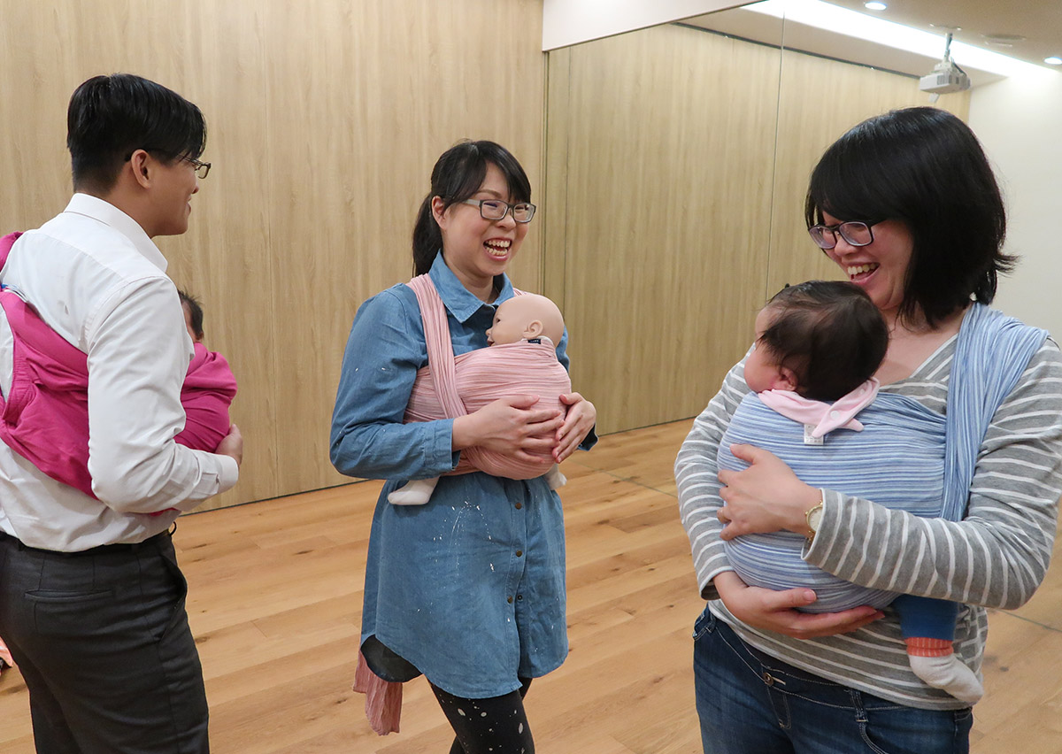 揹巾課程中家長們互相交流與歡笑，爸爸也積極參與其中。這顯示 Oovi 的課程不僅教授綁法技巧，更提供新手父母重要的心理支持 (Emotional Support) 與社交圈，讓育兒路不孤單。