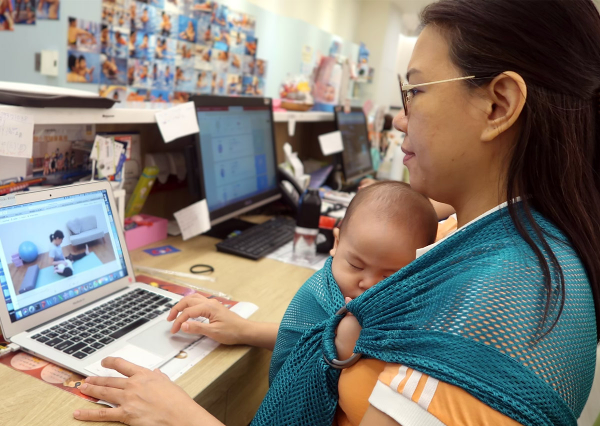 媽媽使用透氣揹巾在電腦前工作同時安撫熟睡寶寶。這張照片完美詮釋揹巾穿戴如何幫助家長達成工作與育兒平衡 (Work-Life Balance)，透過解放雙手讓在家工作 (WFH) 變得更從容。