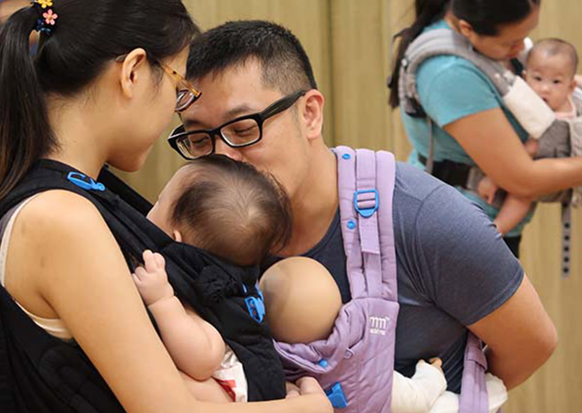 爸爸使用教具娃娃練習揹巾操作，媽媽揹著寶寶親吻額頭，展現共同育兒 (Co-parenting) 的美好畫面，強調揹巾穿戴是促進父嬰連結 (Father-Infant Bonding) 的最佳工具。