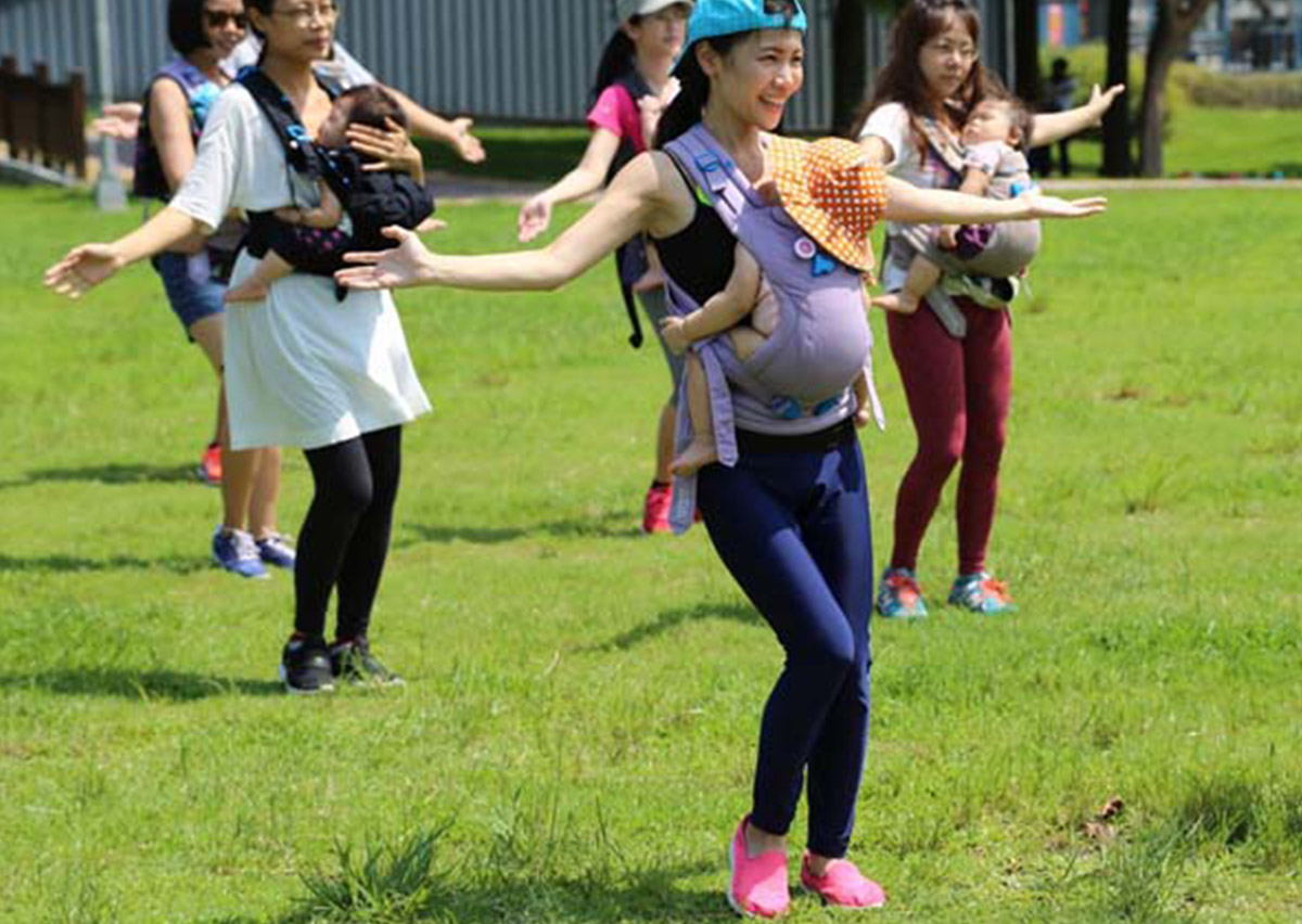 媽媽們在戶外草地上揹著寶寶進行揹巾律動 (Babywearing Dance)，透過伸展動作找回產後核心力量與身體平衡，同時讓寶寶在媽媽懷中享受大自然的感官刺激。