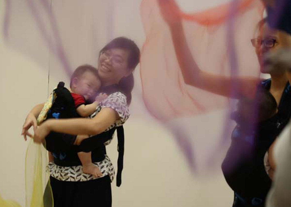 媽媽揹著寶寶舞動彩色絲巾，結合音樂律動與視覺追蹤訓練，這是一種結合感覺統合 (Sensory Integration) 的創意親子互動，增進寶寶對色彩與空間的認知。