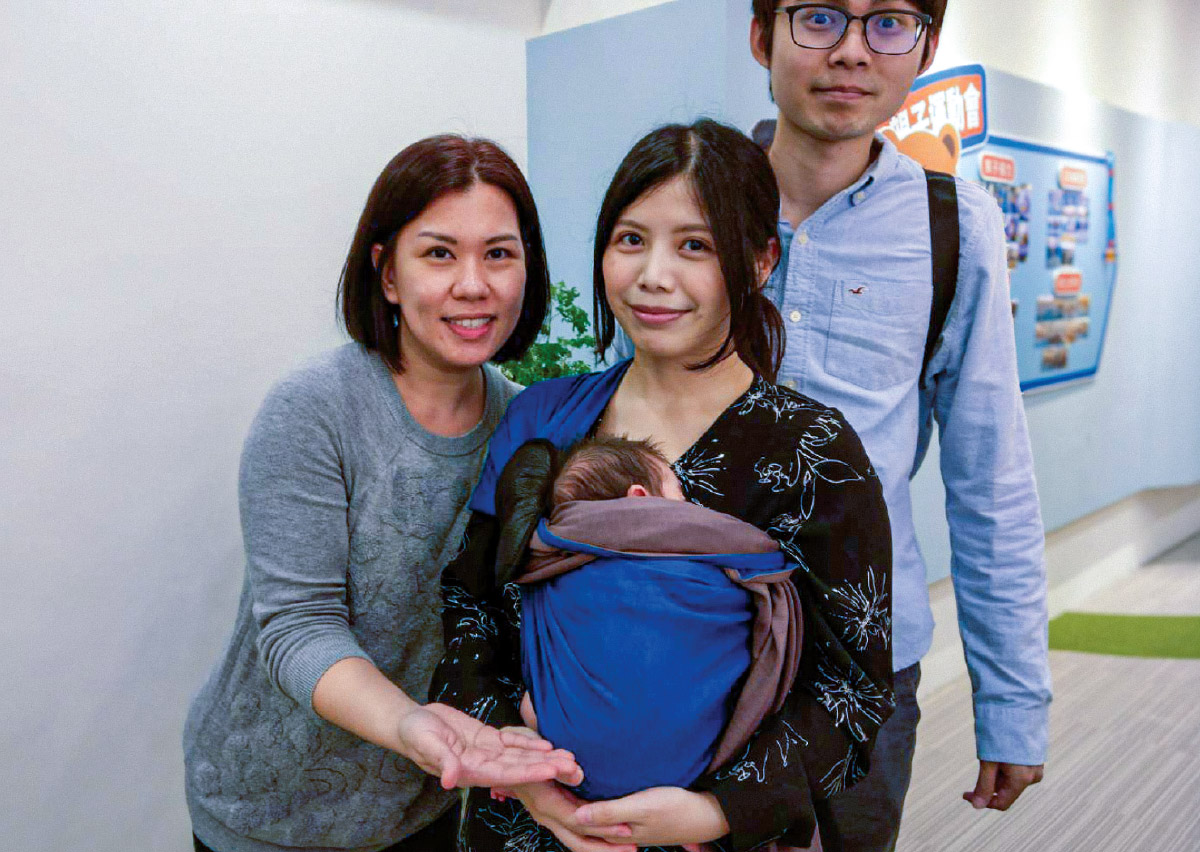 新手爸媽與 Oovi 顧問合影，媽媽使用長布揹巾將新生兒安穩揹在胸前，展示透過專業諮詢選擇合適揹巾，能有效降低新手父母焦慮，讓育兒生活更自信、從容。