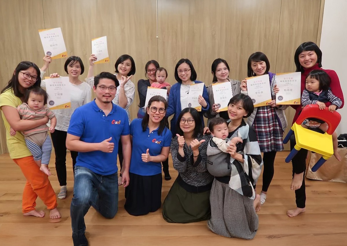 Oovi 揹巾穿戴5.C.M.師資培訓結業合影：見證專業與溫度的起點。多位女性學員手持國際認證證書，懷中抱著嬰幼兒，展現結訓的喜悅與專業成果。前方身著 Oovi 藍色制服的講師比出讚的手勢，背景為木質溫馨教室。