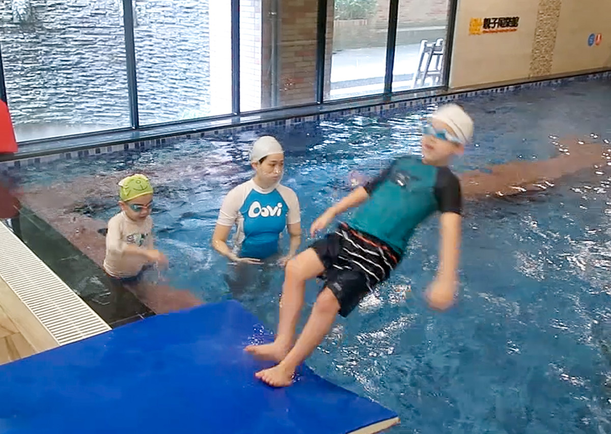 一名幼兒從跳水墊背後躍入溫水泳池，教練在旁貼身守護，展示水域安全教育中培養勇氣與身體協調的關鍵時刻。