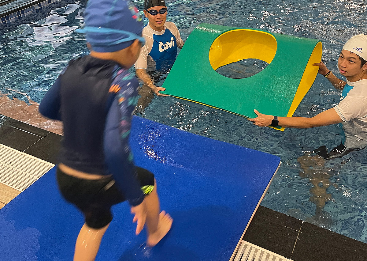 兩位教練在水中舉起圓環浮板，引導孩子精準跳入環中，結合爆發力與目標瞄準的高階體能整合訓練。