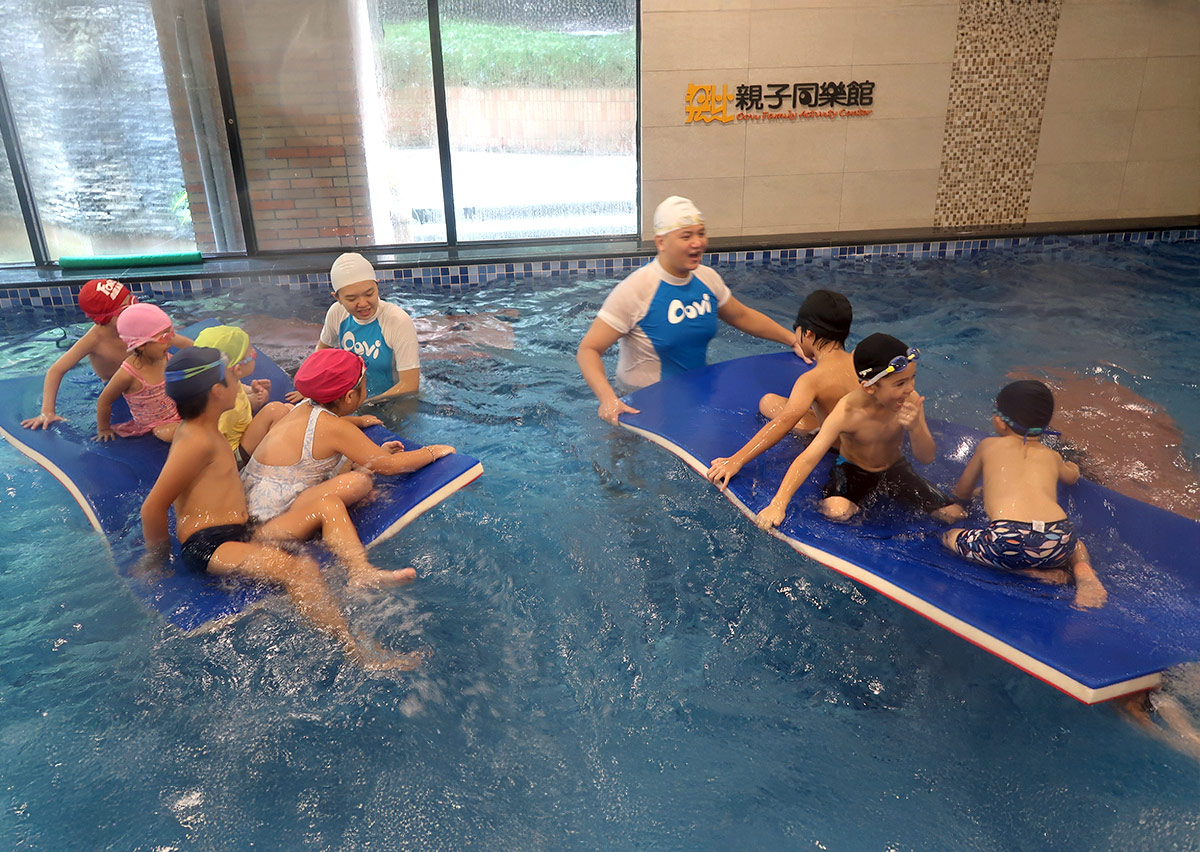 多名孩童分別坐在大型藍色浮板上，在教練引導下練習群體平衡，結合趣味競賽與社交互動。