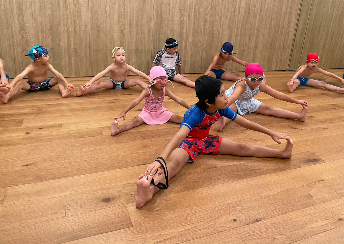 一群孩童在木地板教室進行坐姿分腿伸展，透過正確姿勢預防運動傷害，並提升關節活動度與肌肉彈性。