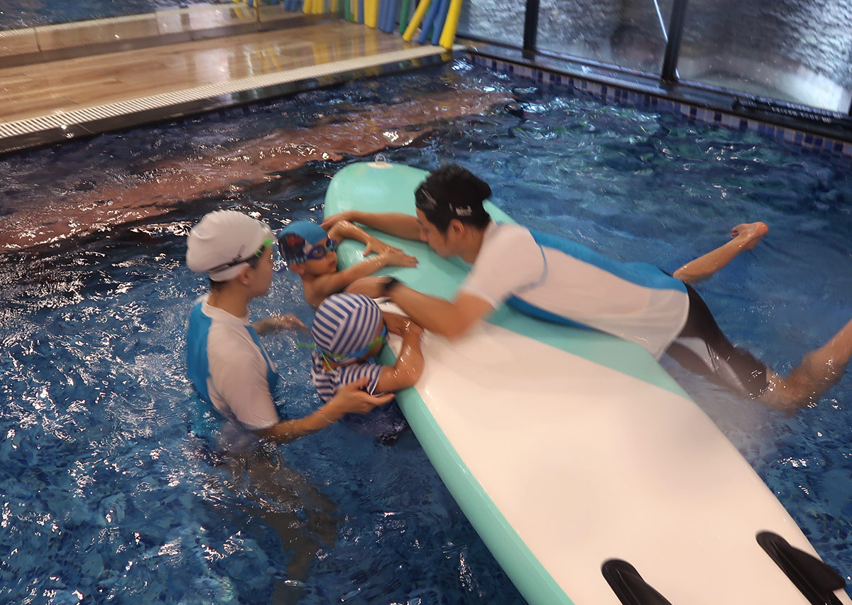 孩童與教練在超大型立槳板（SUP）旁互動，學習在不穩定水面維持團體平衡與安全自救。