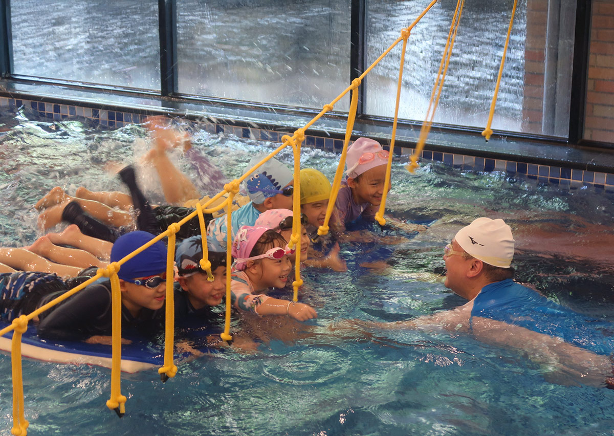 多位孩童趴在大型浮板上，在教練引導下進行水中拍水與漂浮練習，營造放鬆且充滿樂趣的團體學習環境。