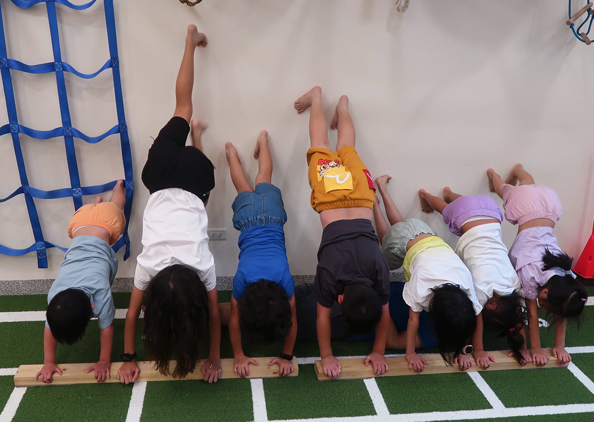 一群孩童在教練指導下進行靠牆手撐訓練，強化肩膀、手臂與核心肌群的負重能力與空間定位感。