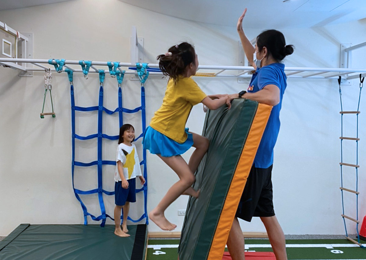 孩童在 Oovi 暑假營隊中挑戰攀爬跳躍動作，並與教練高舉擊掌，從體能遊戲中建立成就感與自信。