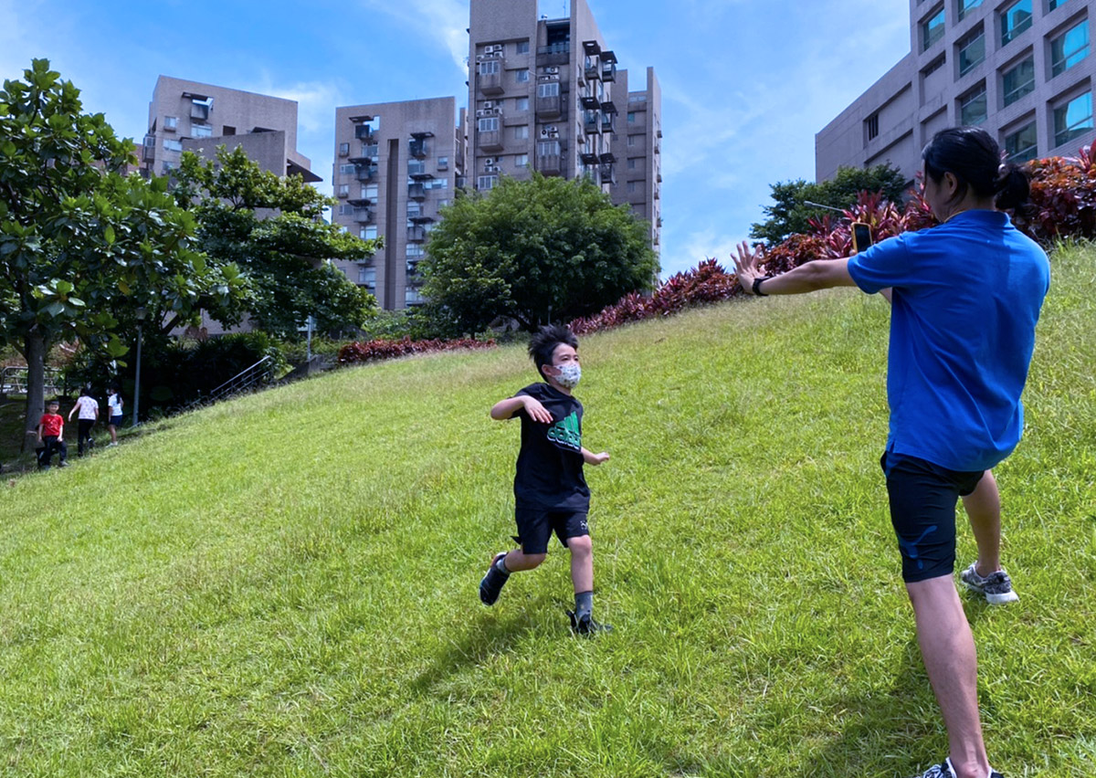 孩童在 Oovi 暑假營隊的戶外綠地向教練奔跑挑戰坡度，透過自然地形的移動訓練鍛鍊下肢力量、心肺耐力與環境適應力。