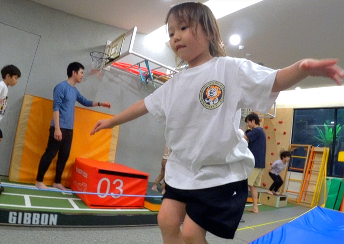 女童在室內體能教室專注地走在 Slackline (走繩) 上,這項極具挑戰性的動態平衡運動能深度訓練核心肌群、腳踝穩定度與專注力 (Focus)。背景展示了 Oovi FMS 課程多元的攀爬與跳躍設施。