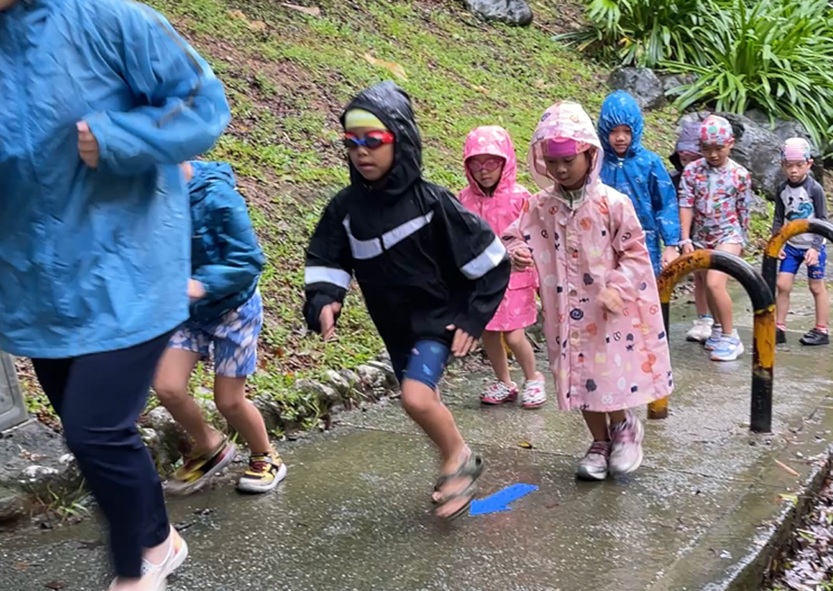 孩子們穿著雨衣在雨中進行戶外路跑 挑戰,跟隨地面箭頭指引前進。這不僅鍛鍊心肺耐力,更培養孩子面對惡劣天氣的環境適應力與堅持到底的韌性 (Resilience)。
