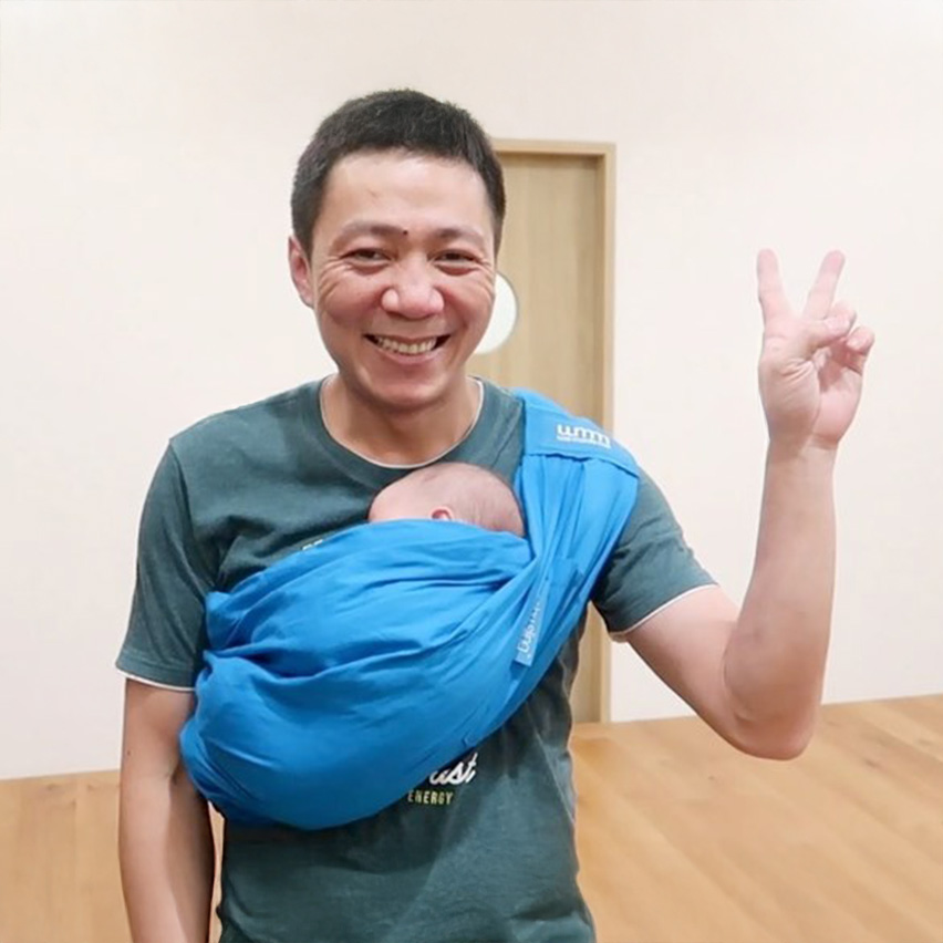 爸爸試穿藍色單肩揹巾後開心比出勝利手勢，展示這種款式操作簡易且穿脫快速的特性，非常適合爸爸短程外出或居家安撫使用，提升育兒成就感。