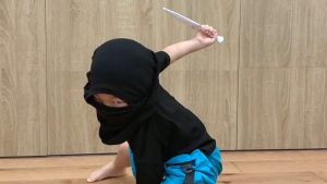 一名參與 Oovi 小忍者訓練營隊的小朋友穿著黑色忍者蒙面頭巾，正做出帥氣的忍者蹲姿並手持紙製道具。背景為教室的木質牆面。這類沉浸式角色扮演活動旨在透過趣味化教學，幫助孩子在遊戲過程中建立高度專注力、肢體控制能力與自我認同感。