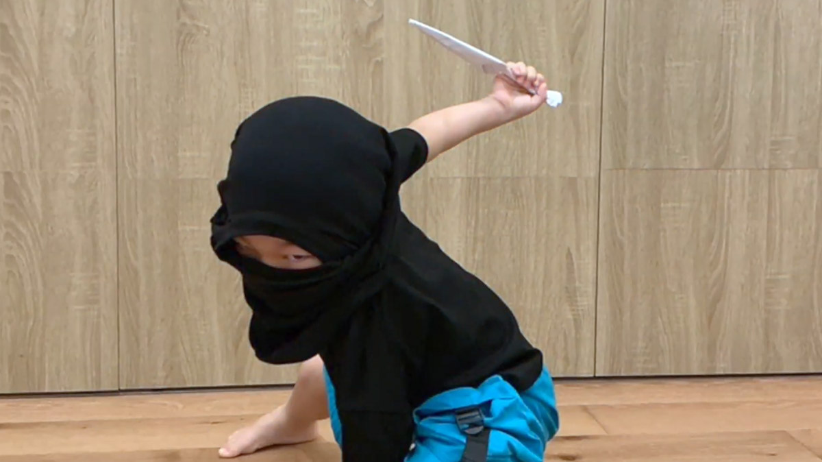 一名參與 Oovi 小忍者訓練營隊的小朋友穿著黑色忍者蒙面頭巾，正做出帥氣的忍者蹲姿並手持紙製道具。背景為教室的木質牆面。這類沉浸式角色扮演活動旨在透過趣味化教學，幫助孩子在遊戲過程中建立高度專注力、肢體控制能力與自我認同感。